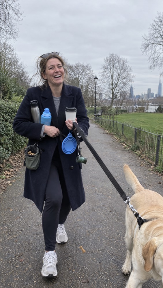 A laughing woman on a park walk juggling a coffee, two water bottles, a clip-on bowl, a treat pouch and a Labrador on a lead